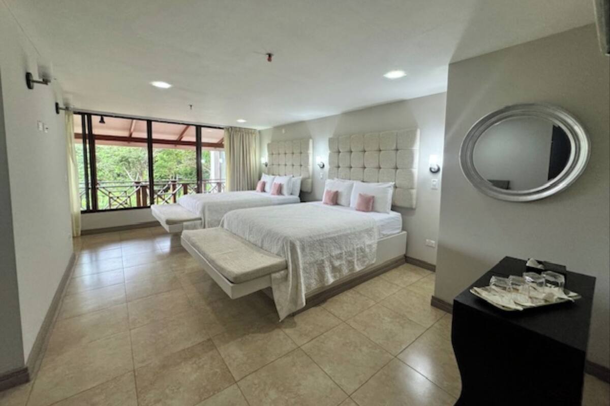A hotel room with two beds, a bench, a round mirror, and a small table with glasses.