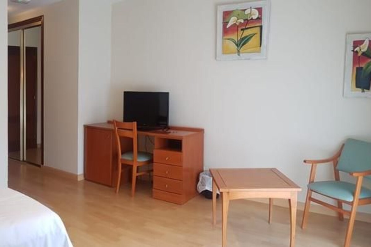 A hotel room with a bed, a desk with a TV, a chair, and a small table.