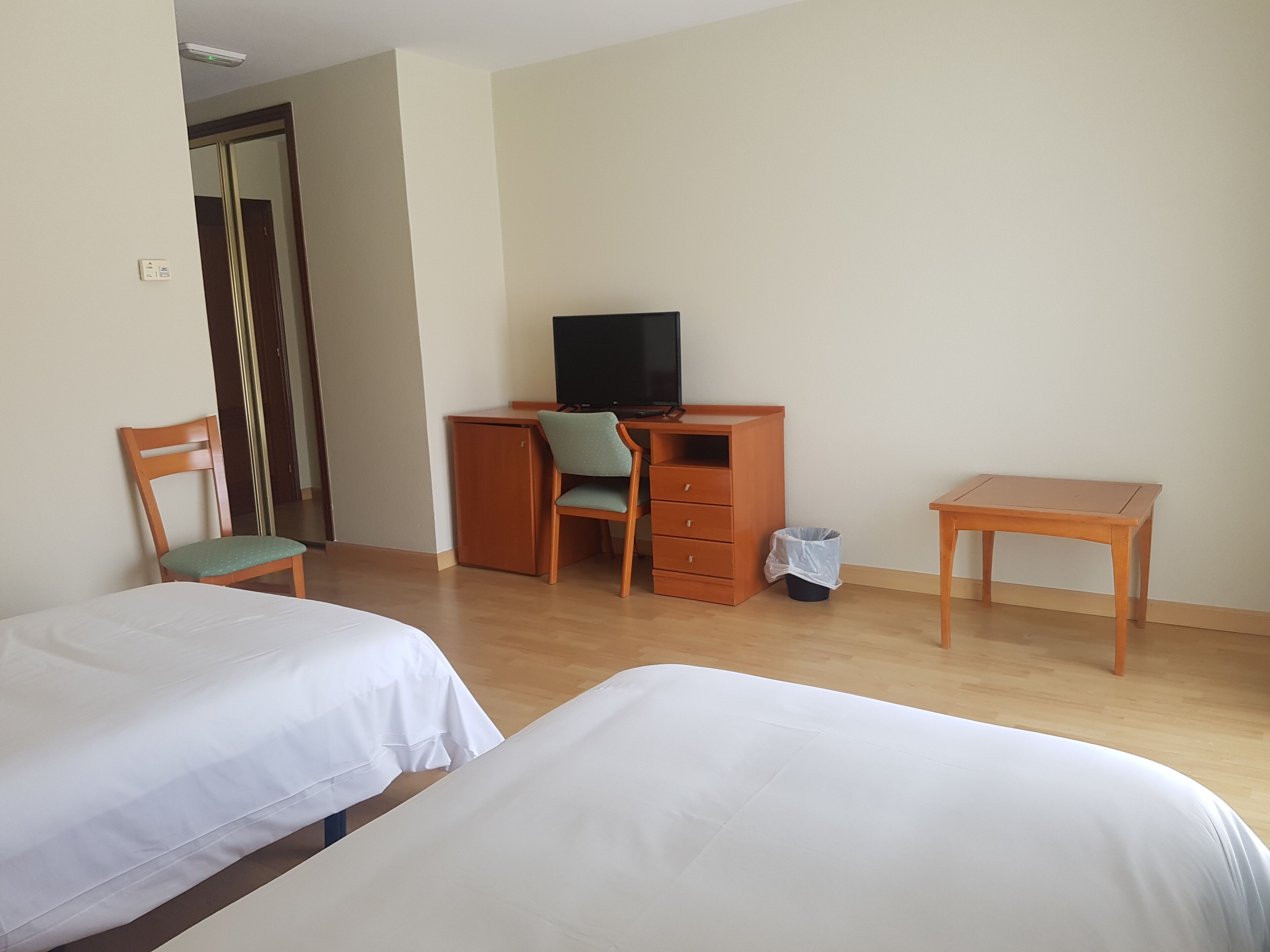 Habitación de hotel con dos camas, un escritorio con silla, un televisor y un pequeño cubo de basura.