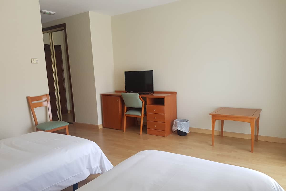Habitación de hotel con dos camas, un escritorio con silla, un televisor y un pequeño cubo de basura.
