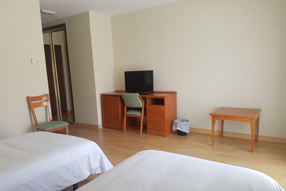 A hotel room with two beds, a desk with a chair, a television, and a small trash bin.