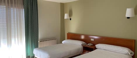A hotel room with two single beds, a wooden headboard, a bedside table with a lamp, and a window with sheer curtains.