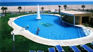 Une piscine agrémentée d’une fontaine centrale, entourée de chaises longues et de palmiers, avec une plage en arrière-plan.