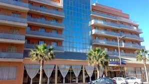 Un hôtel à plusieurs étages, doté d’une tour centrale en verre, de balcons à chaque étage et de palmiers à l’avant.