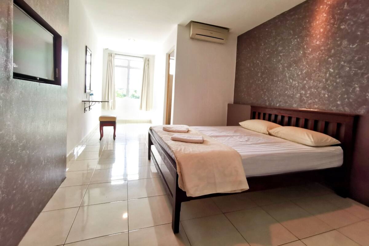 A hotel room with a bed, a bench, a TV, and an air conditioning unit.