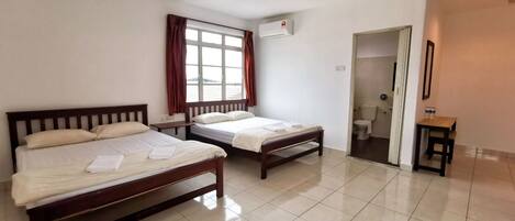 A hotel room with two wooden beds, a window with curtains, an air conditioning unit, a small table, and a door leading to a bathroom.