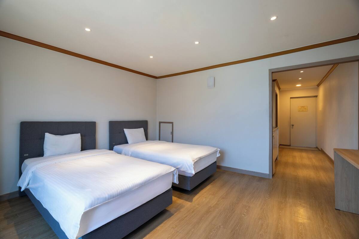 A hotel room with two beds, wooden flooring, and a hallway leading to a door.