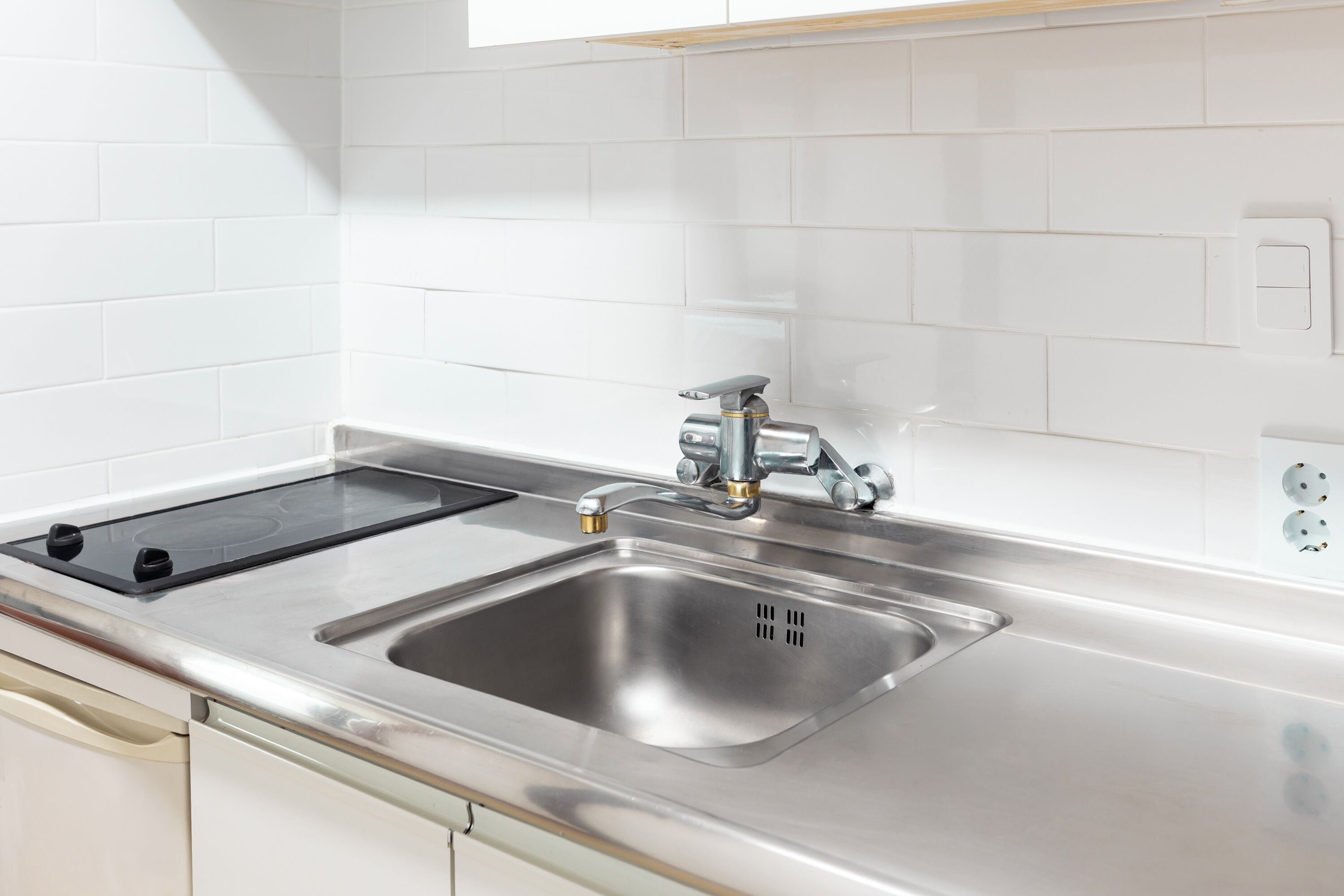 Una cocina con un lavamanos de acero inoxidable, una cocina vitrocerámica negra y paredes cubiertas de azulejos blancos.