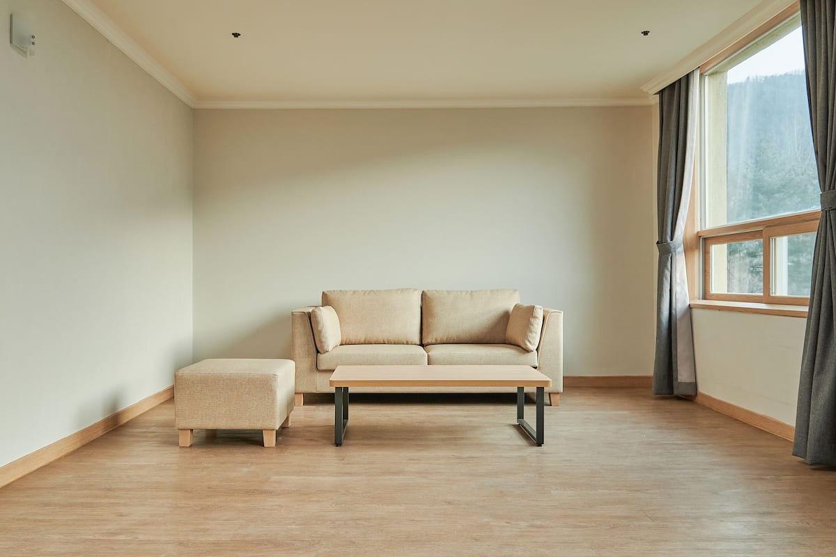A minimalist living room with a sofa and ottoman, a large window with curtains, and a light wooden floor.