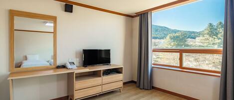 Habitación de hotel con escritorio de madera, televisor de pantalla plana, espejo y ventana grande con vistas a un área boscosa.