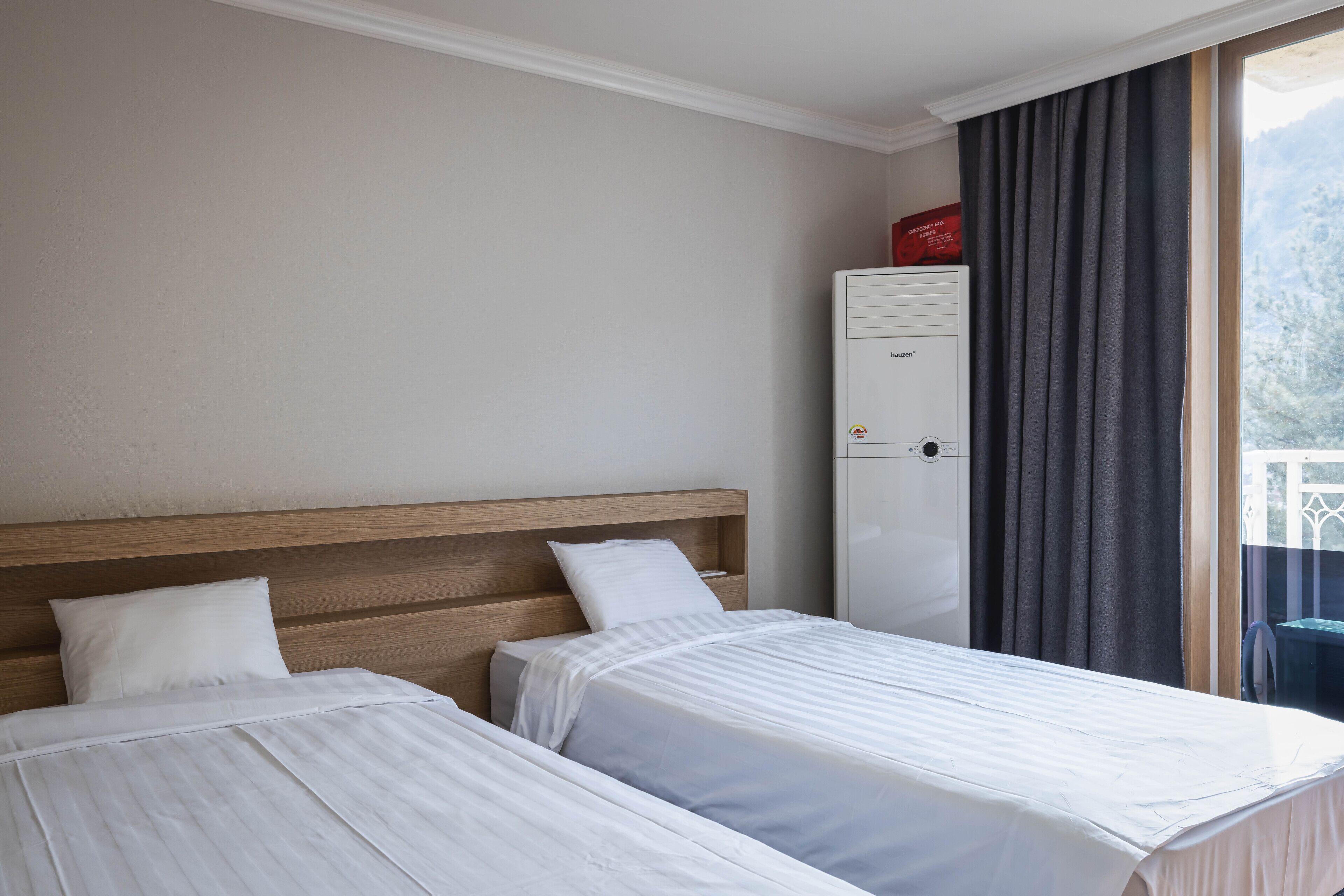 A hotel room with two single beds, a wooden headboard, a white wardrobe, and a view of a balcony with curtains.
