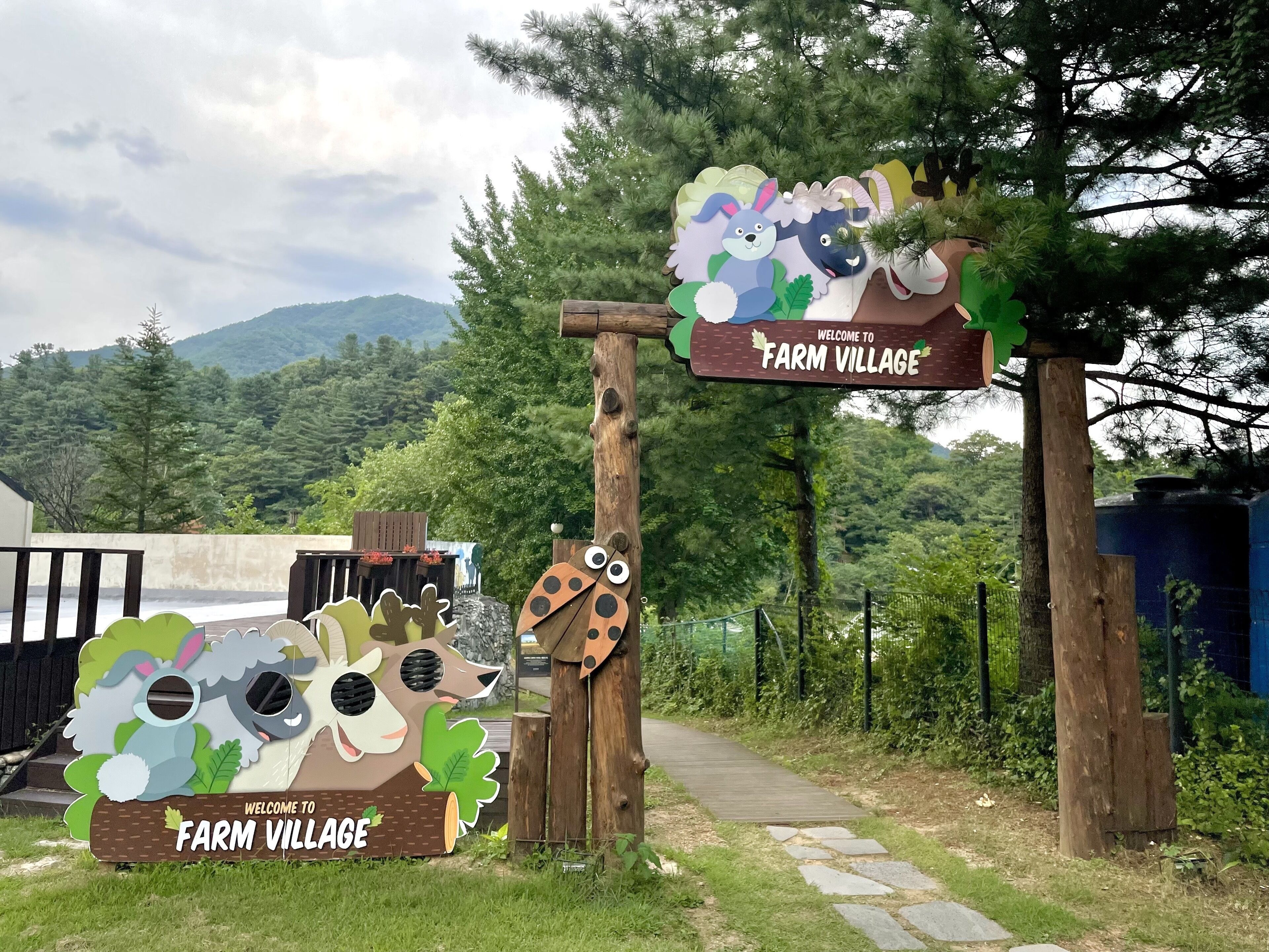 Ein hölzernes Wegweiser mit der Aufschrift 'Welcome to Farm Village' und einem Pfad, der in einen bewaldeten Bereich führt.