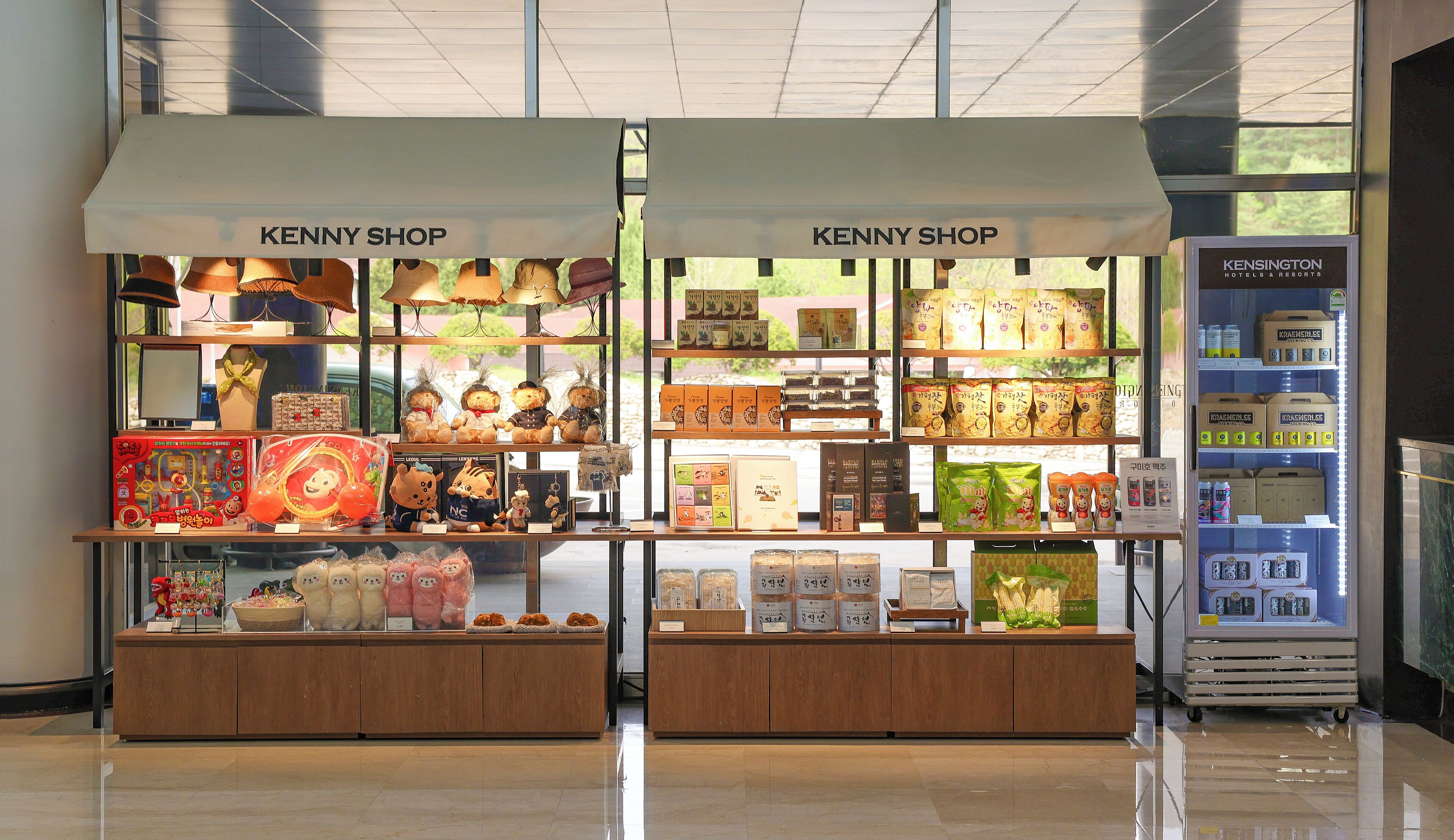 Two KENNY SHOP kiosks with various merchandise, including plush toys and packaged goods, in a well-lit indoor setting.