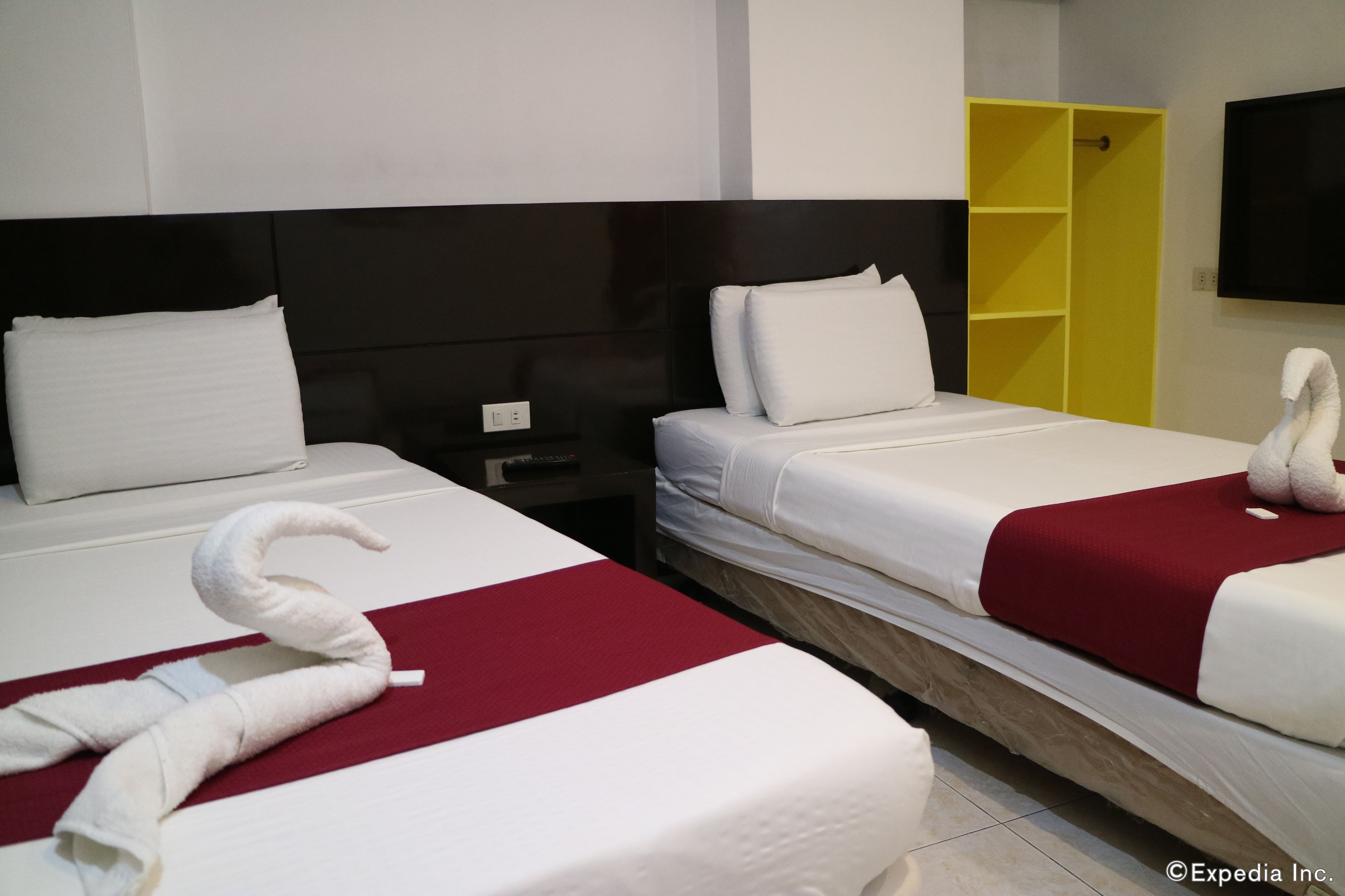 A hotel room with two beds, each with white linens and red accents, a yellow wardrobe, and a flat-screen TV mounted on the wall.