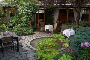 A stone-paved outdoor seating area with wicker furniture and tables, surrounded by lush greenery and a wooden structure. - American House Baletowa (Warsaw)