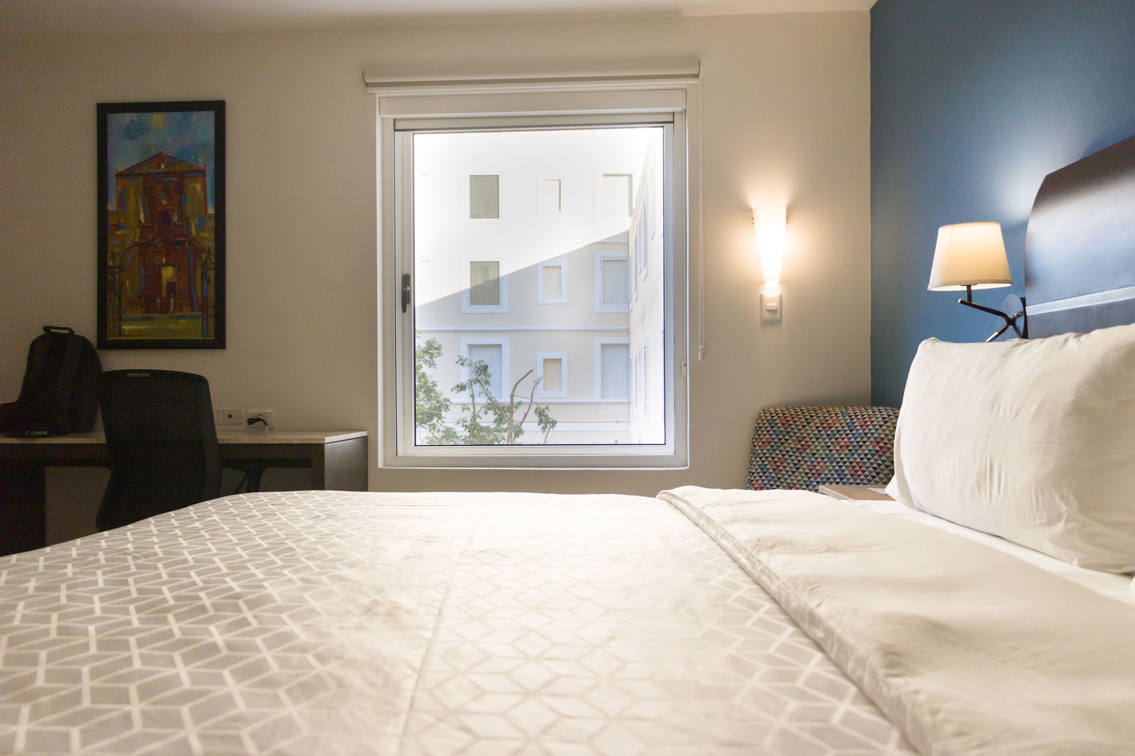 A hotel room with a bed, a desk, a chair, a window with a view of buildings, and a wall-mounted lamp.