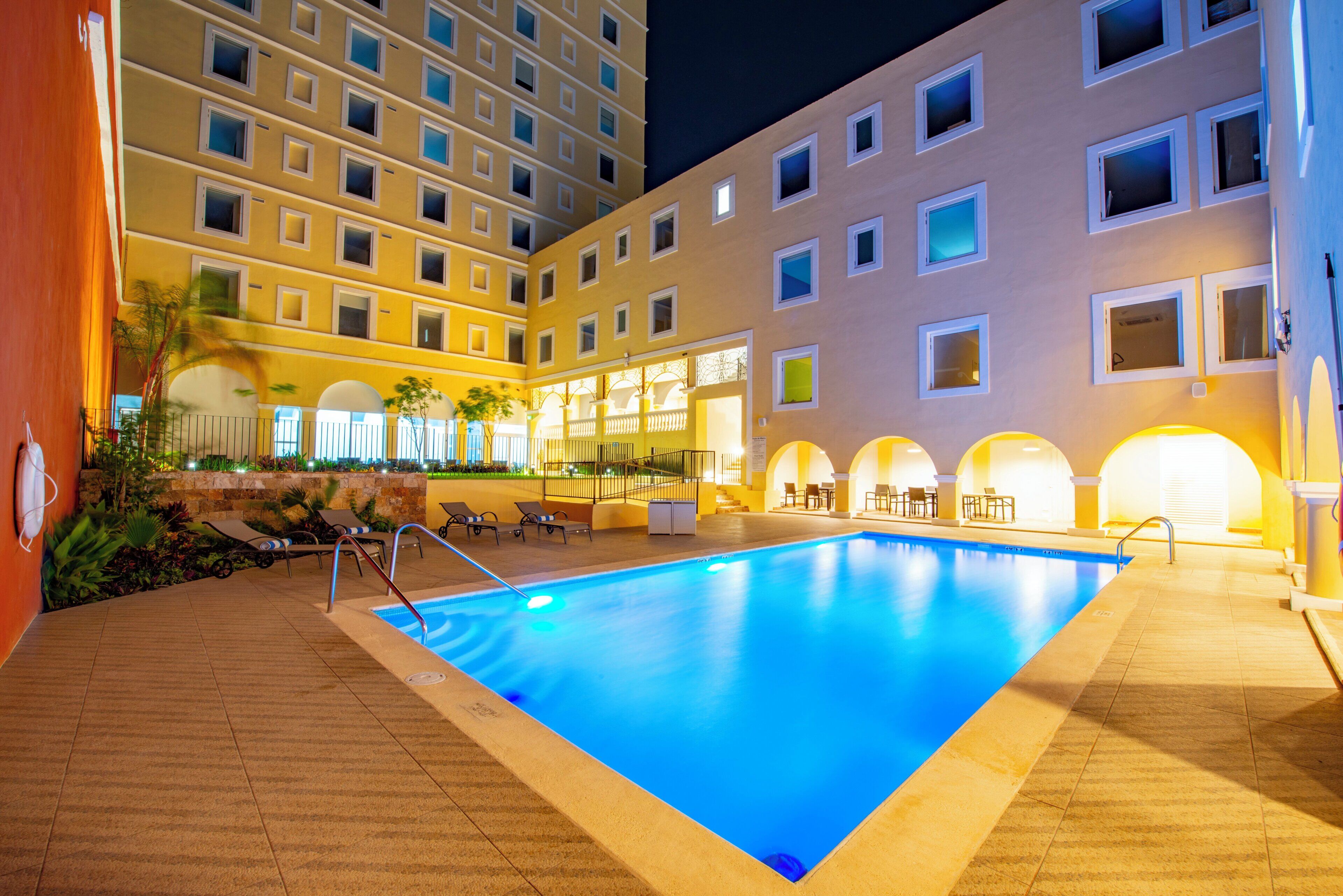 A swimming pool with sun loungers and a poolside bar, surrounded by a multi-story hotel building with balconies and windows.