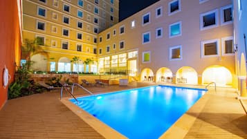A swimming pool with sun loungers and a poolside bar, surrounded by a multi-story hotel building with balconies and windows.