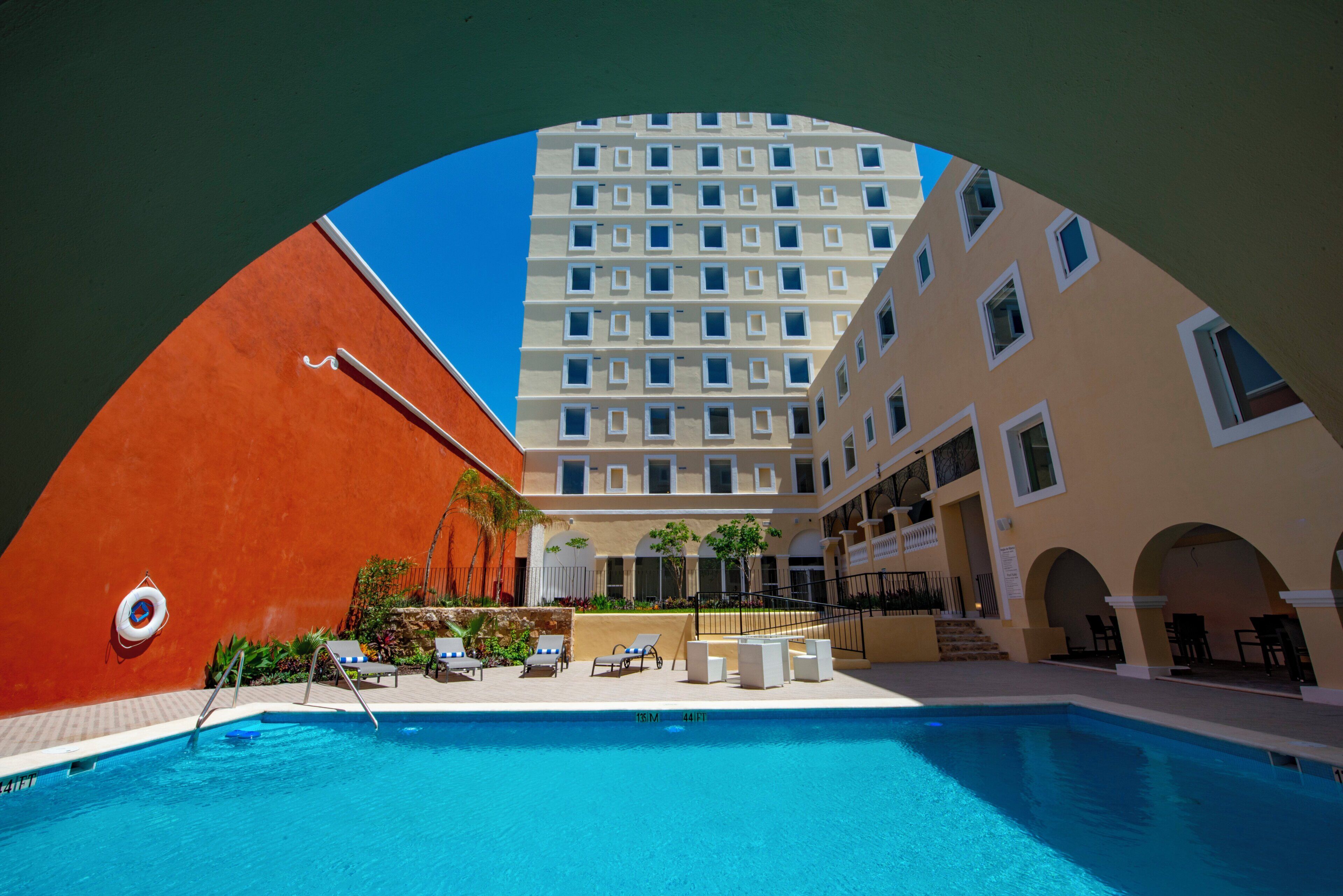 A hotel pool area with a clear view of the pool, lounge chairs, and a building with multiple windows.