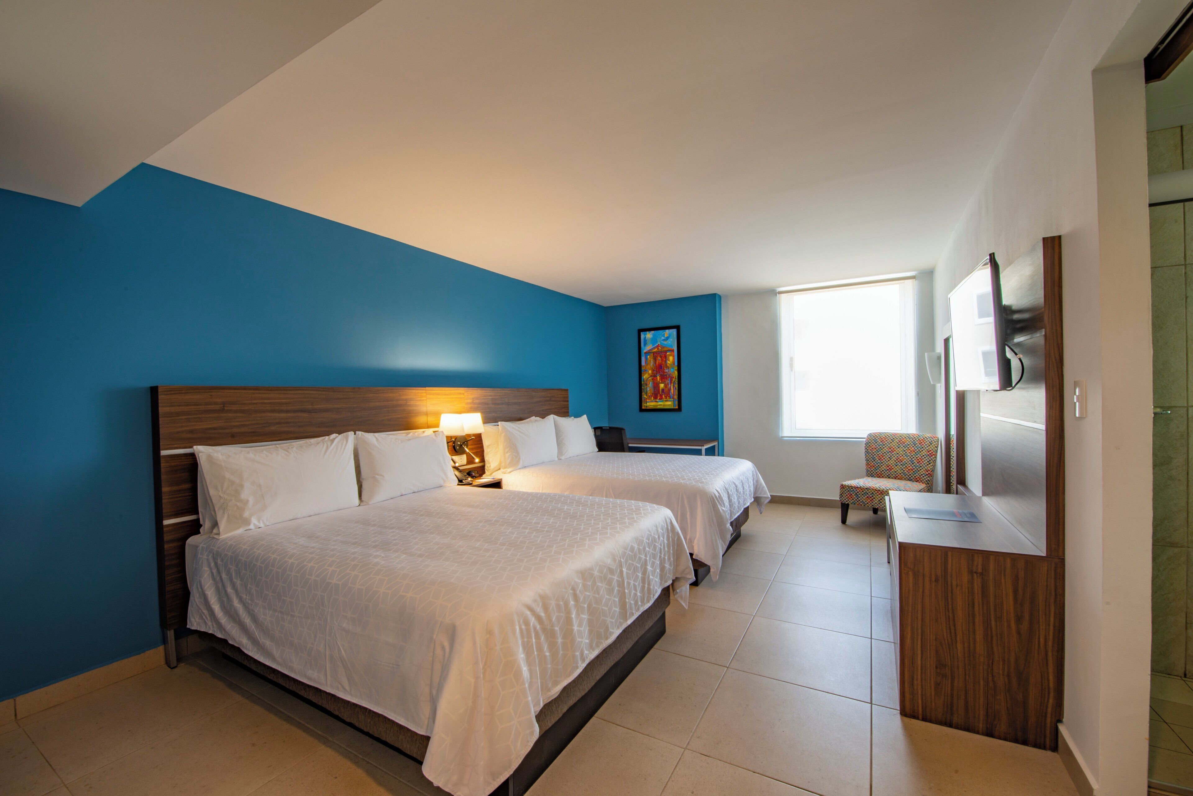 A hotel room with two beds, a desk, a chair, a TV, and a window.