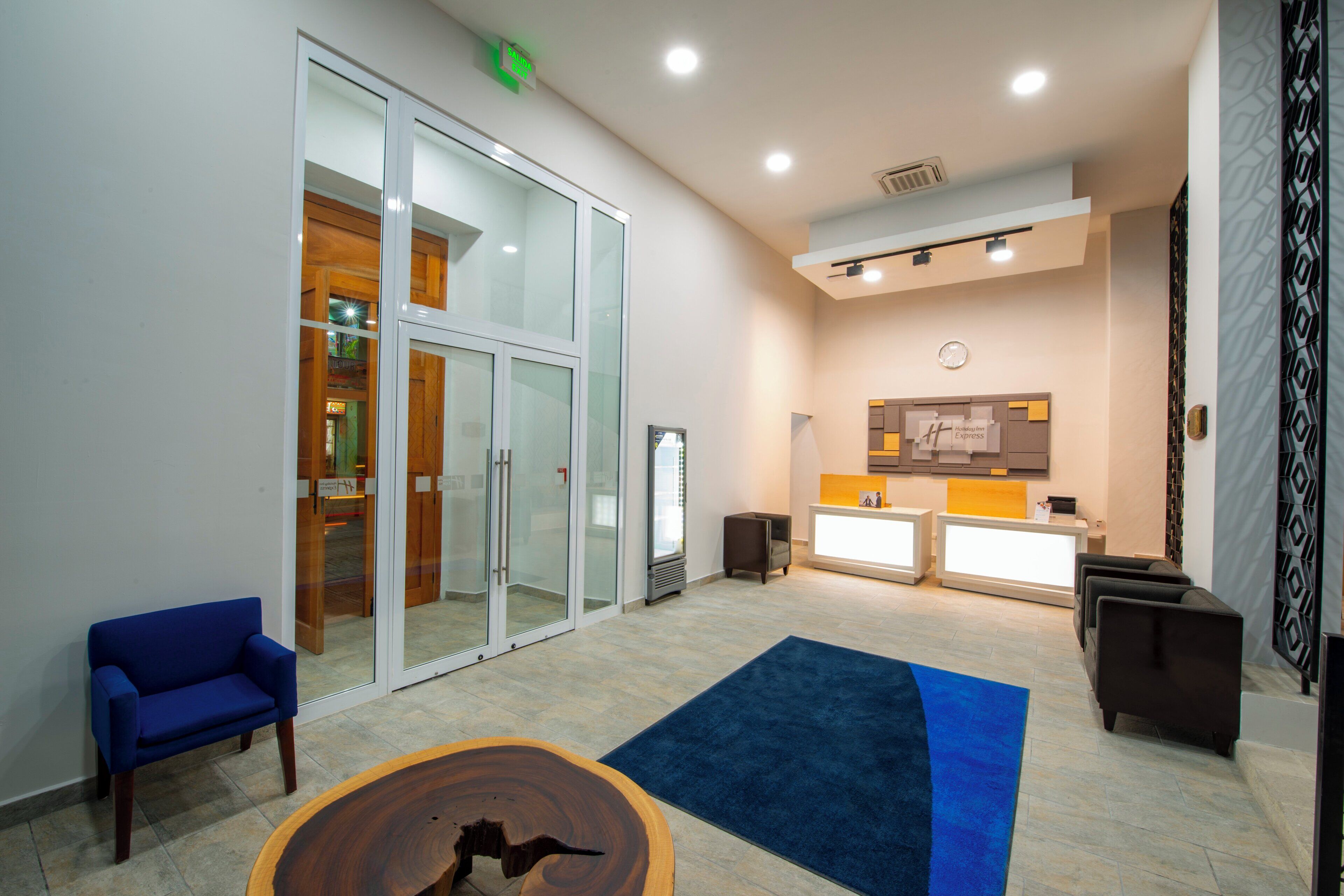 Un vestíbulo de hotel con mostrador de recepción, zona de estar y puertas de vidrio que dan a un pasillo.