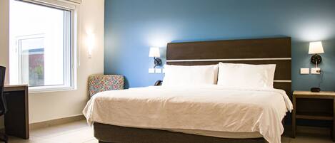 A hotel room with a large bed, a desk, and a chair. There are two wall-mounted lamps and a window with blinds.