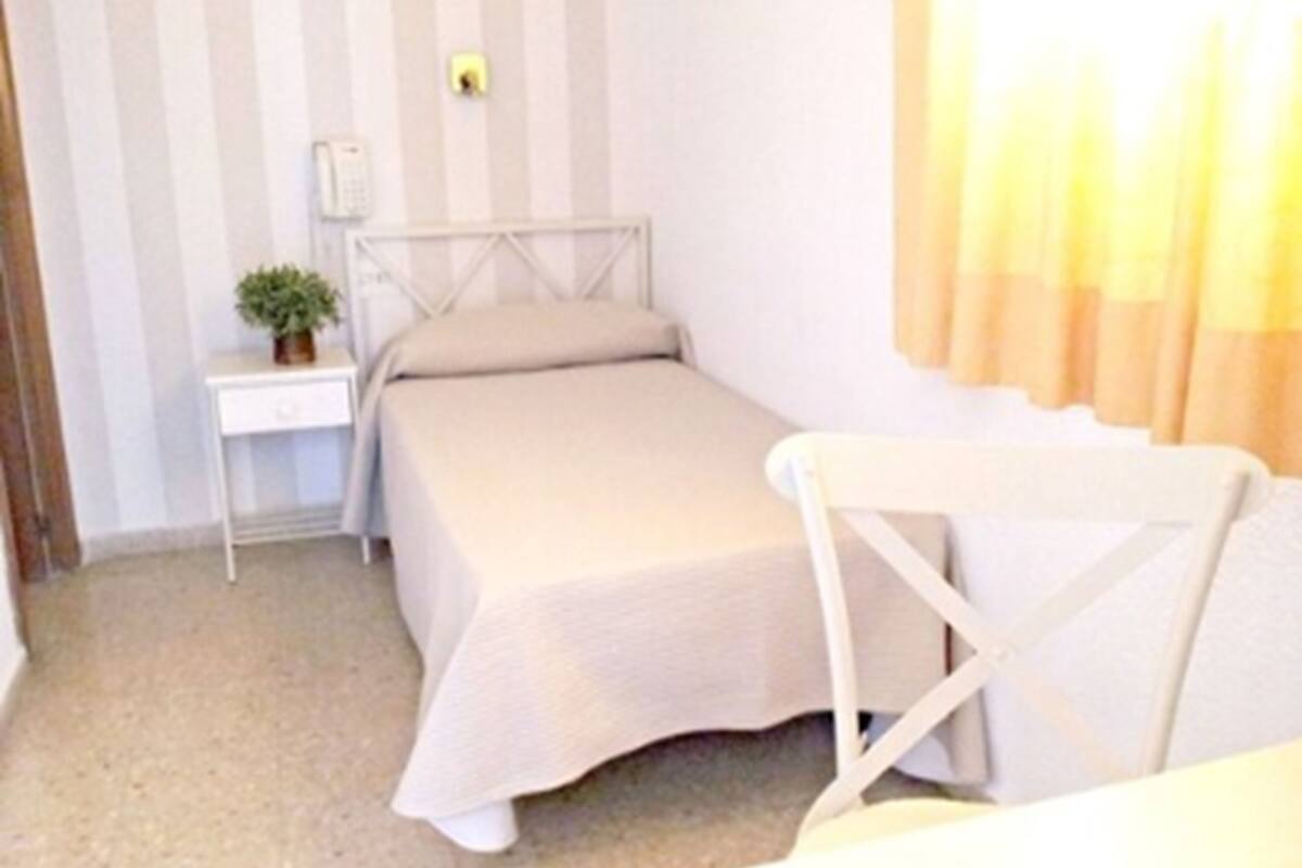 Una habitación pequeña con una cama individual, una mesita de noche, una silla y un control remoto sobre la mesa.