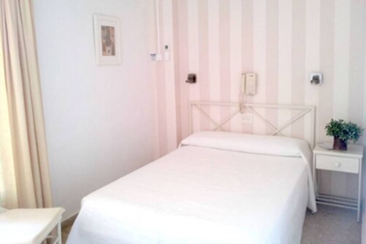 Habitación individual con una cama blanca, una mesita de noche, una pequeña planta en maceta y una pared a rayas.