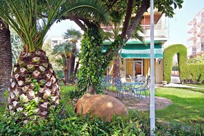 A courtyard with a palm tree, a large rock, and a building with a green awning. - Tramontana (Benicassim)