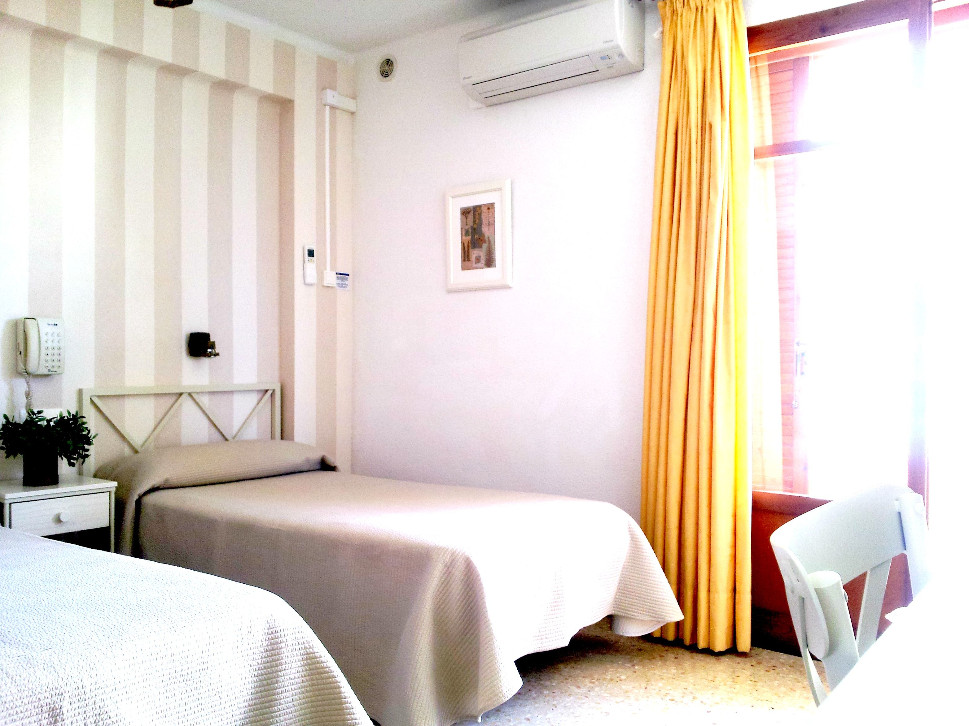 Una camera d'albergo con due letti, un comodino con lampada, un telefono e una piccola pianta.