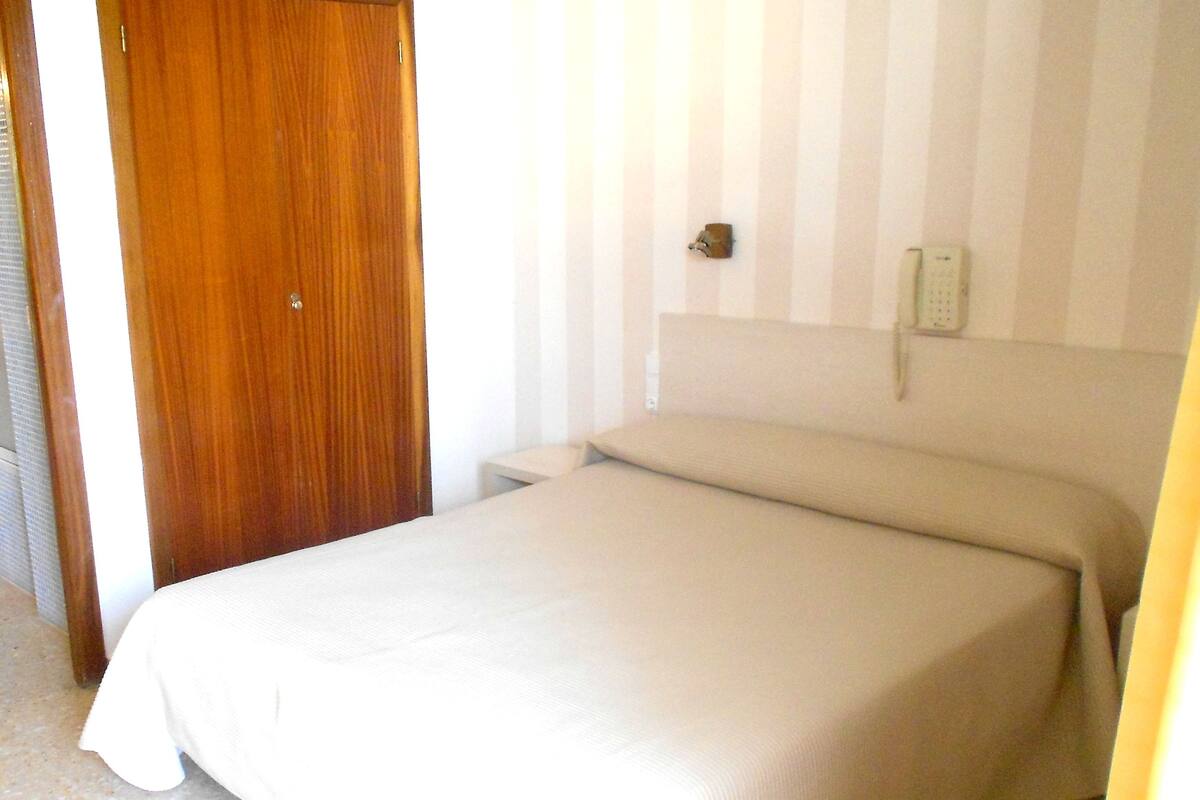Un dormitorio con puerta de madera, una cama con cubrecama color beige, una mesita de noche pequeña y un teléfono instalado en la pared.