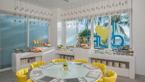 Un espace repas moderne avec une table ronde en verre entourée de chaises jaunes, une grande fenêtre donnant sur des palmiers et un mur décoratif orné d’un panneau avec la lettre « b ».