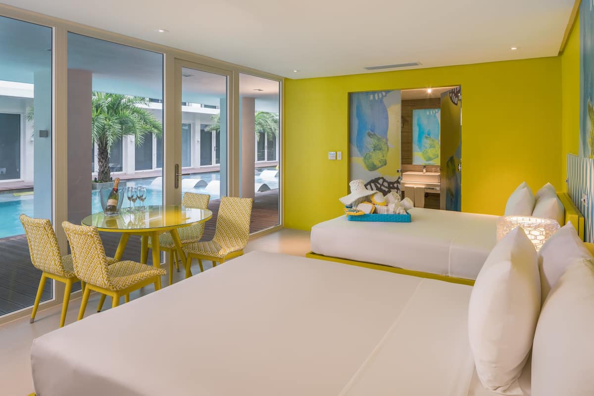 Una habitación de hotel moderna con una cama grande, un comedor con sillas amarillas y vistas a una piscina y palmeras.