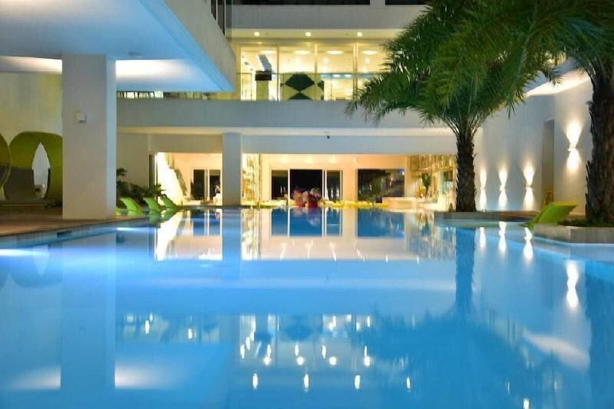 A swimming pool with a reflection, palm trees, and a building with multiple floors and glass railings.