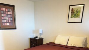 A neatly made bed with a dark red bedspread, a wooden nightstand with a lamp, and a framed artwork on the wall. - Tip Top Motel Cafe & Bakery (Lihue)