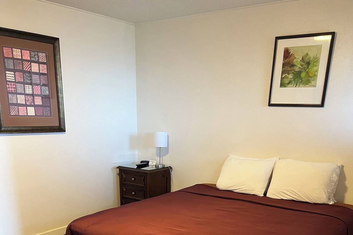 Una cama bien hecha con una colcha rojo oscuro, una mesita de noche de madera con una lámpara y una obra de arte enmarcada en la pared.