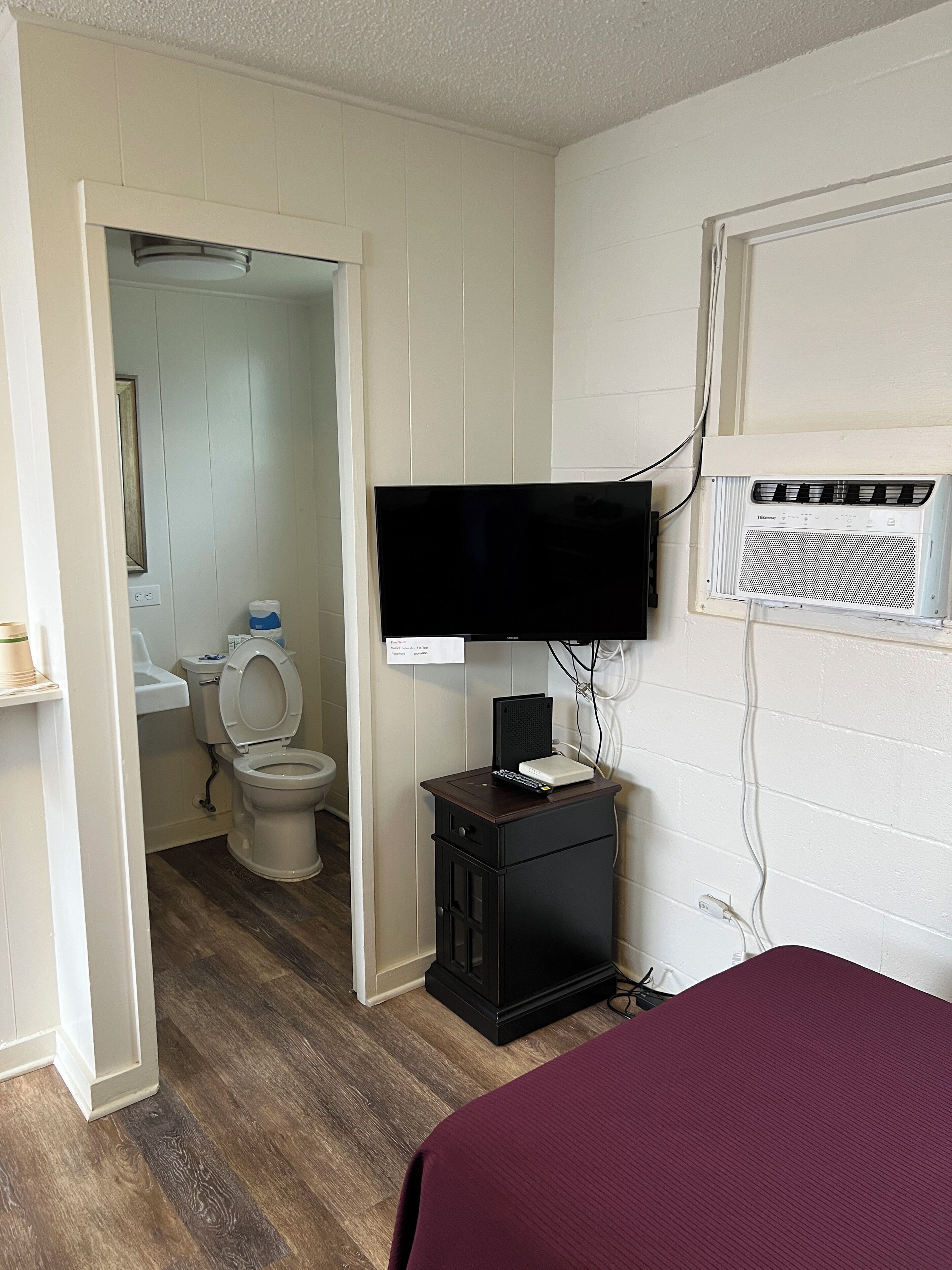 A bedroom with a bed, a TV mounted on the wall, a small wooden cabinet, and a bathroom visible through an open door.