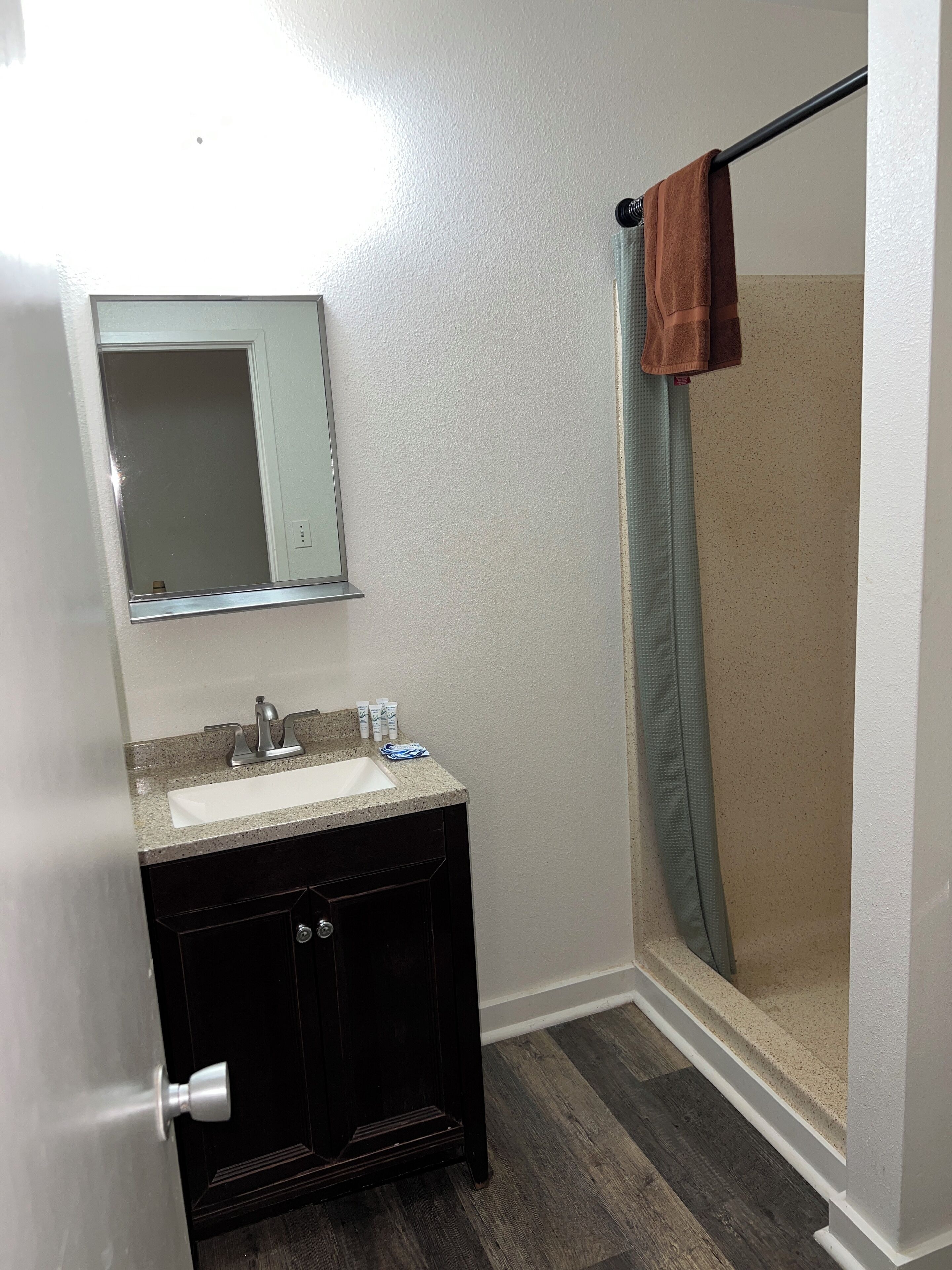 Un baño con lavamanos, espejo, mueble de madera oscura y zona de ducha con una toalla colgada en la puerta.