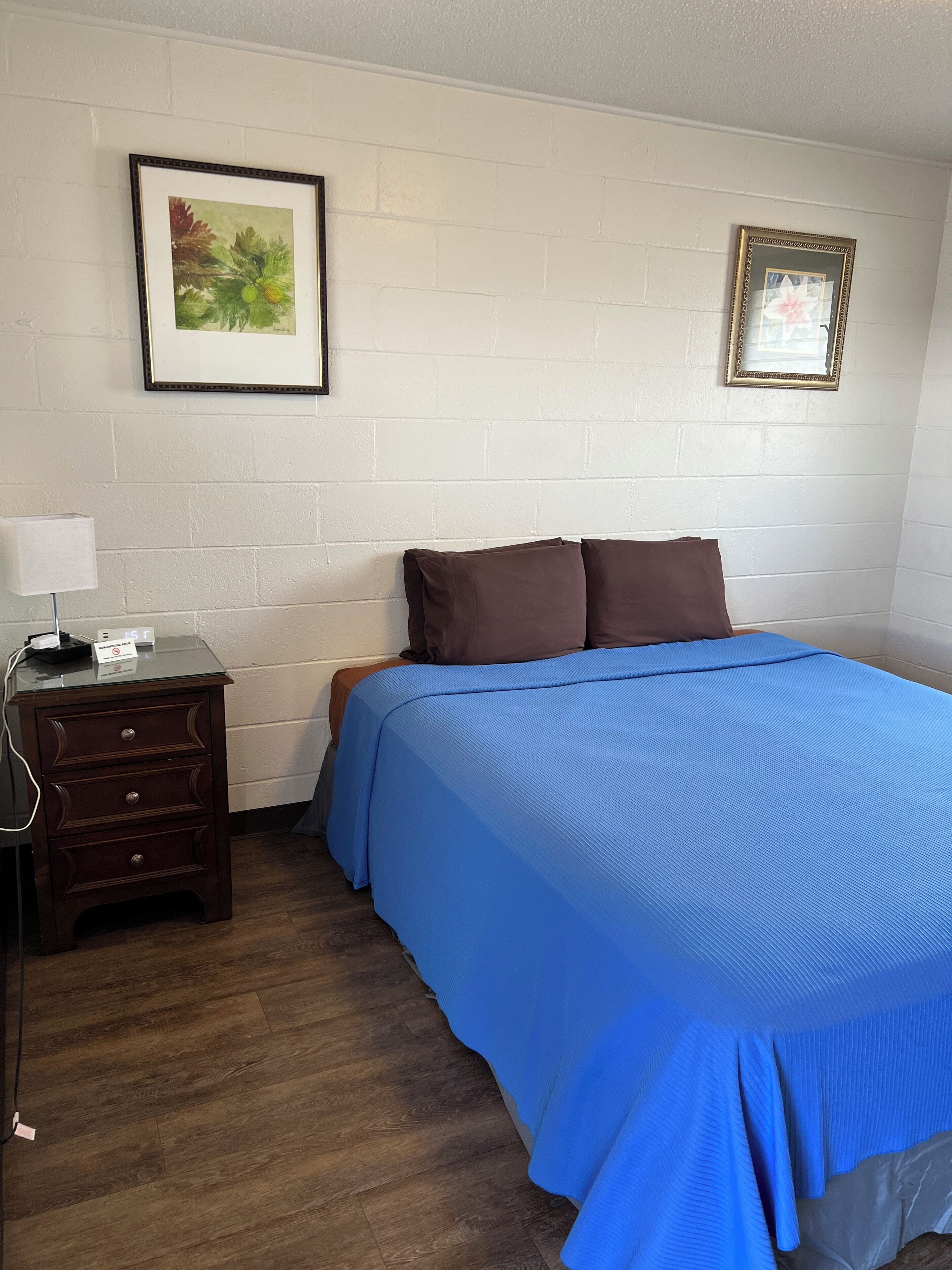 Une chambre avec une couvre-lit bleue, une table de chevet en bois surmontée d’une lampe, et deux tableaux encadrés accrochés au mur.