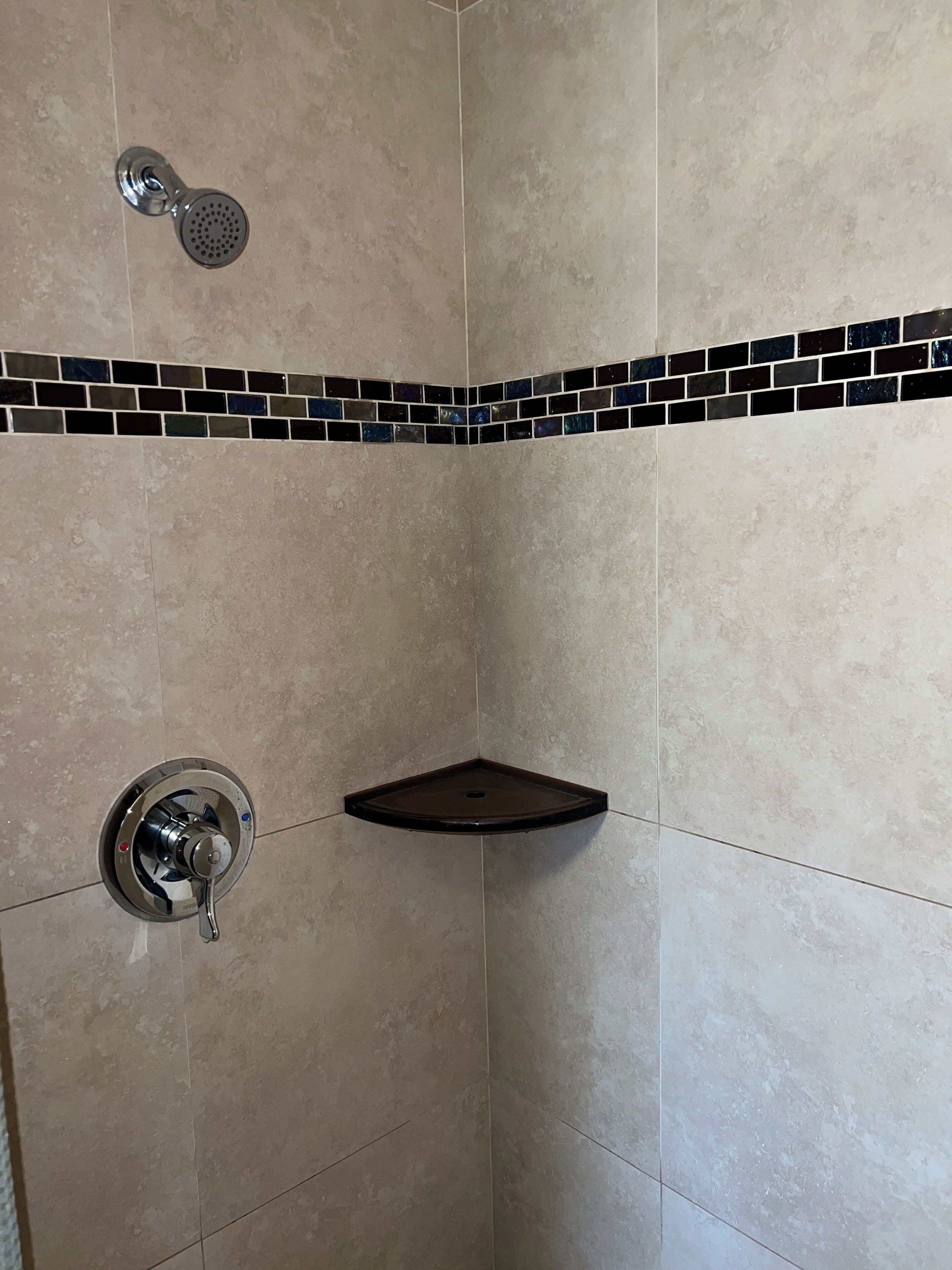 Esquina de un baño con baldosas color crema, un estante negro, un cabezal de ducha y un grifo.