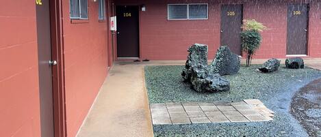 Un edificio con puertas numeradas, un corredor cubierto y un pequeño patio con rocas y un árbol.