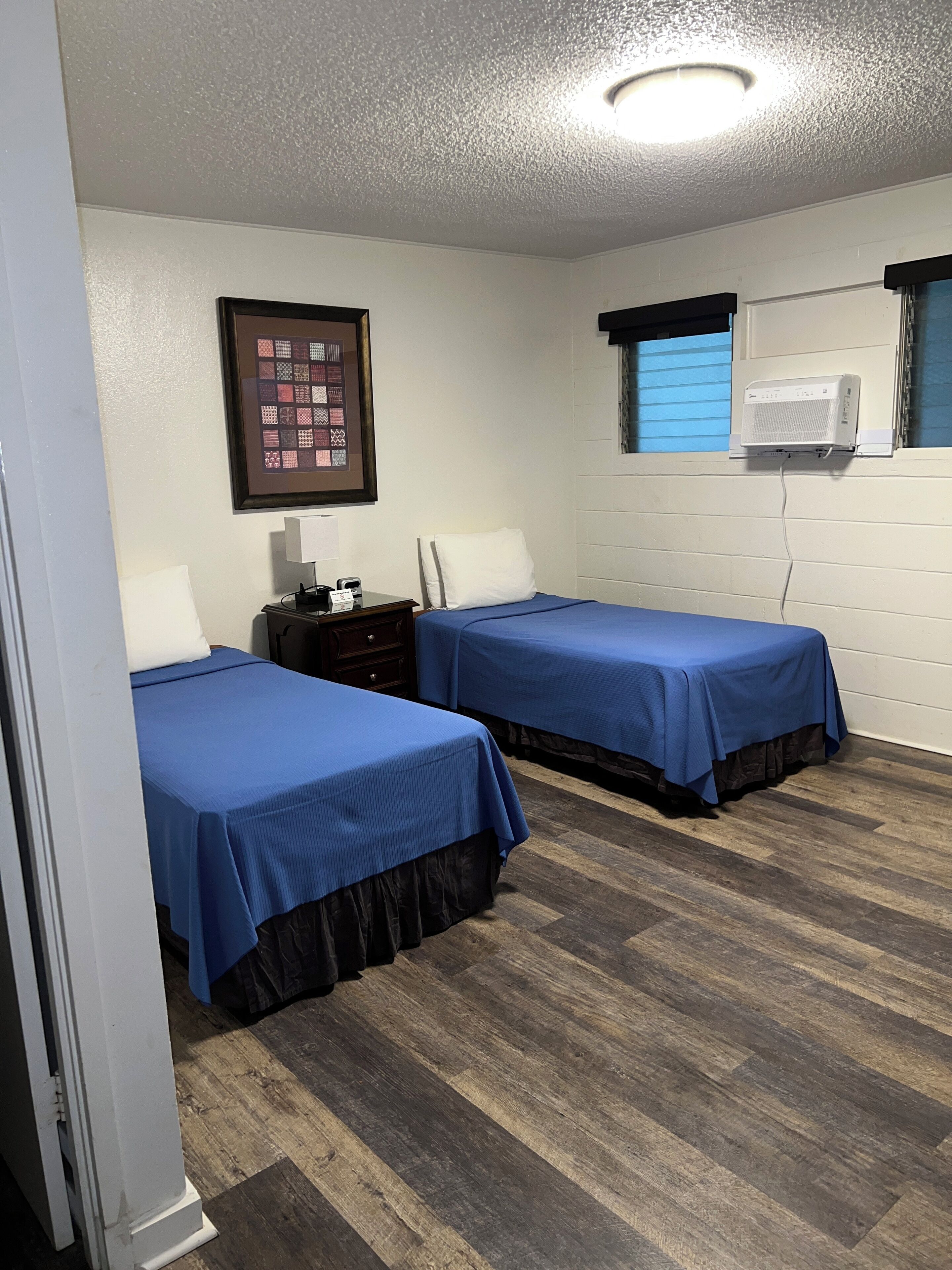 A hotel room with two beds, a nightstand, a framed quilt, a window with blinds, and an air conditioning unit.