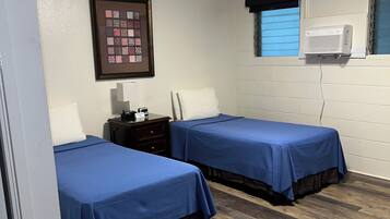 A hotel room with two beds, a nightstand, a framed quilt, a window with blinds, and an air conditioning unit.