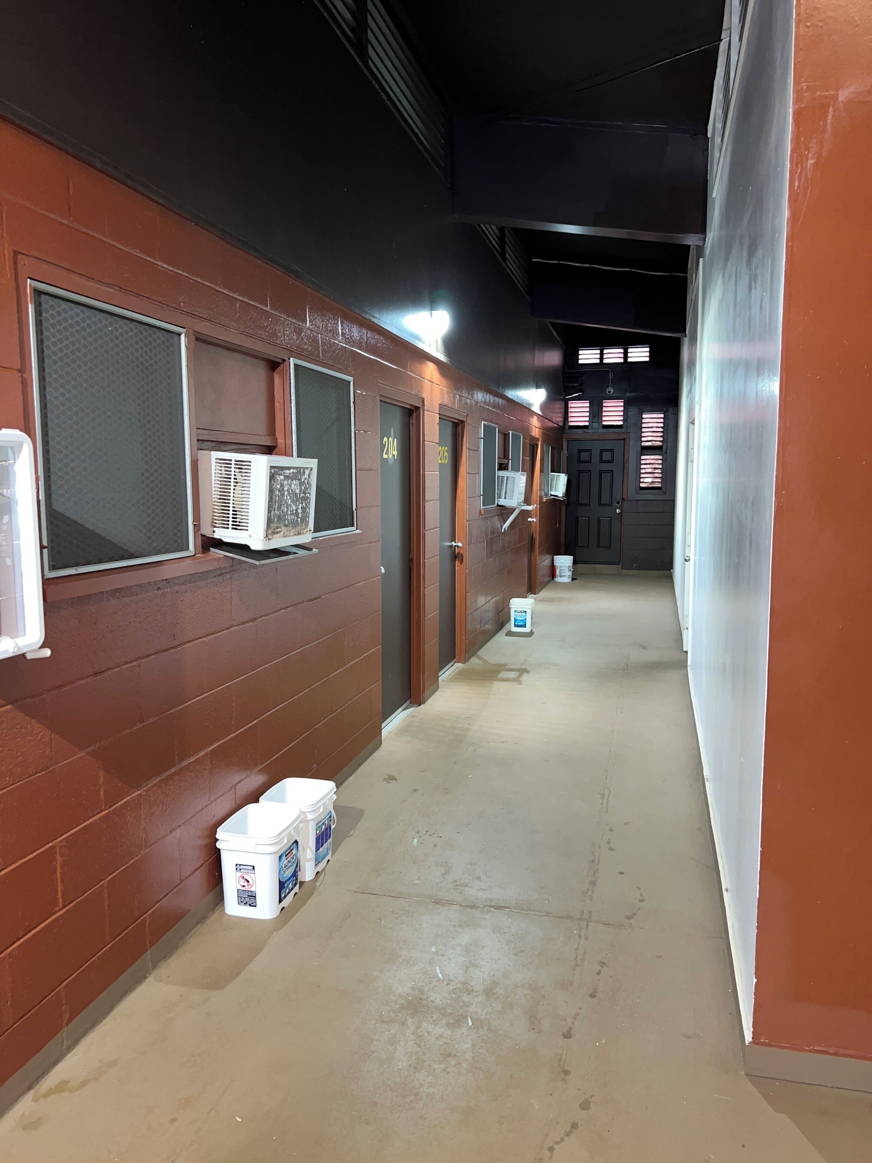 A corridor with numbered doors, air conditioning units, and storage bins.