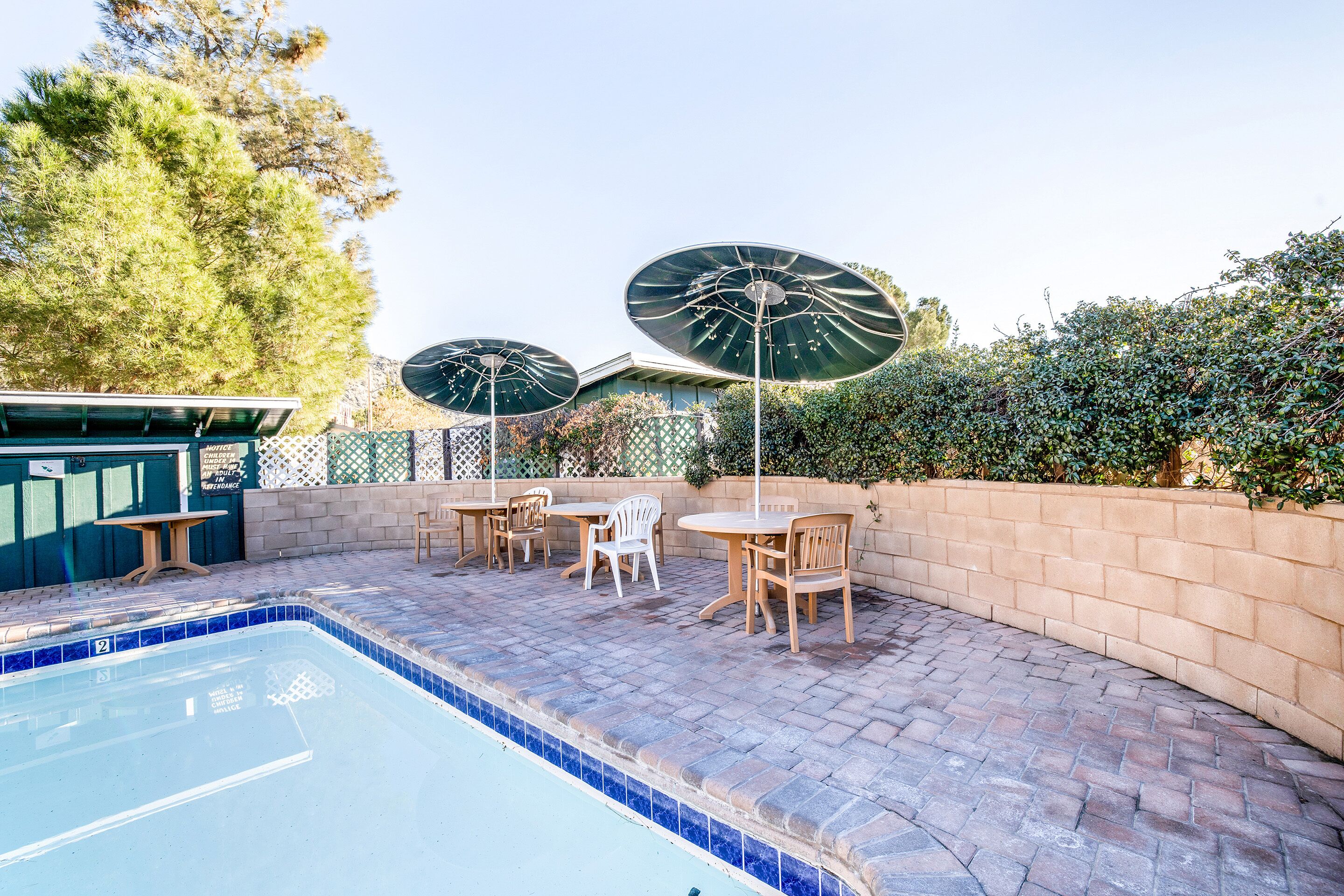 Una piscina con bordes de azulejos, rodeada por un patio de ladrillo, sillas de madera y dos grandes sombrillas.