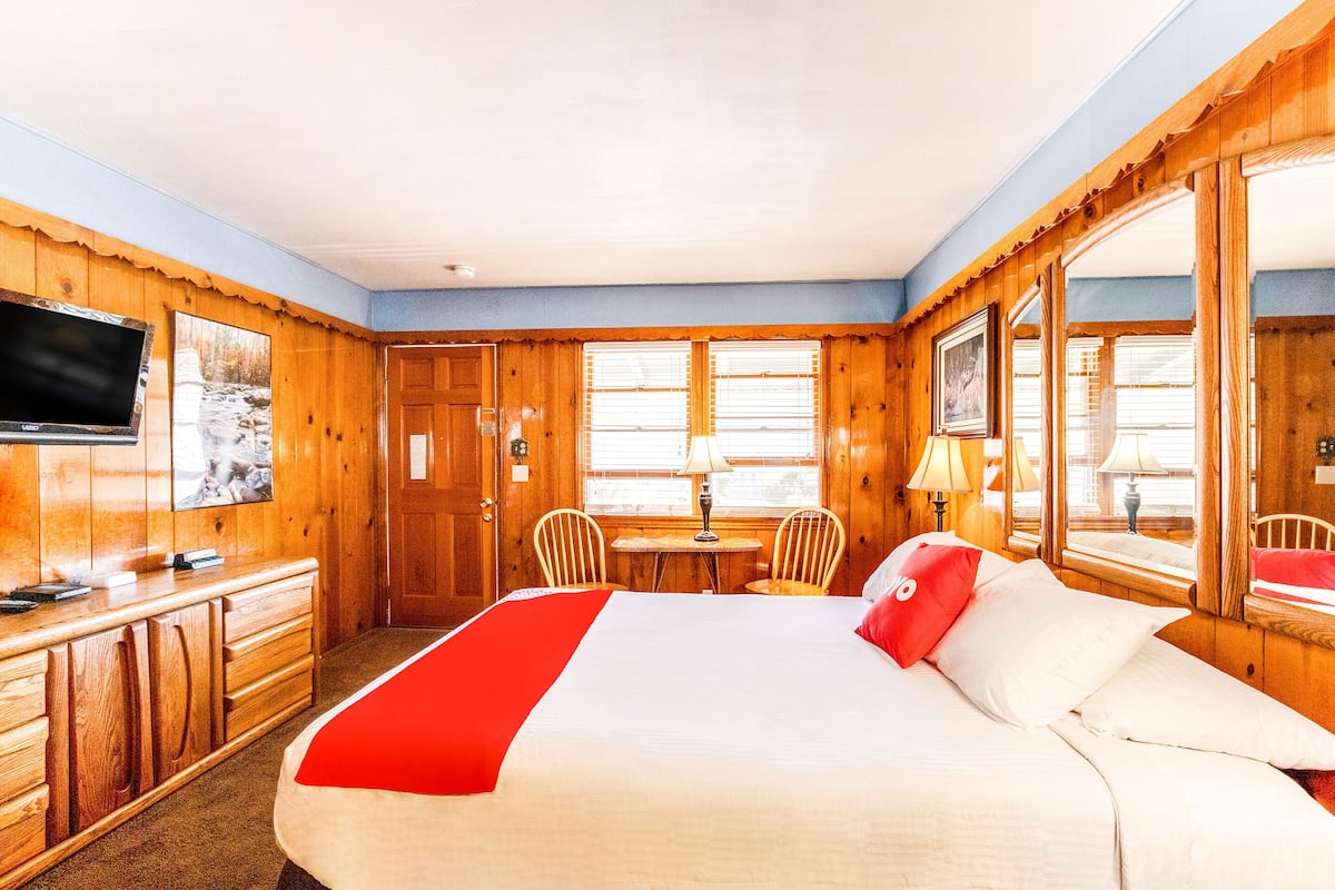 A hotel room with a large bed, wooden walls, a television, a desk with a chair, and a window with blinds.