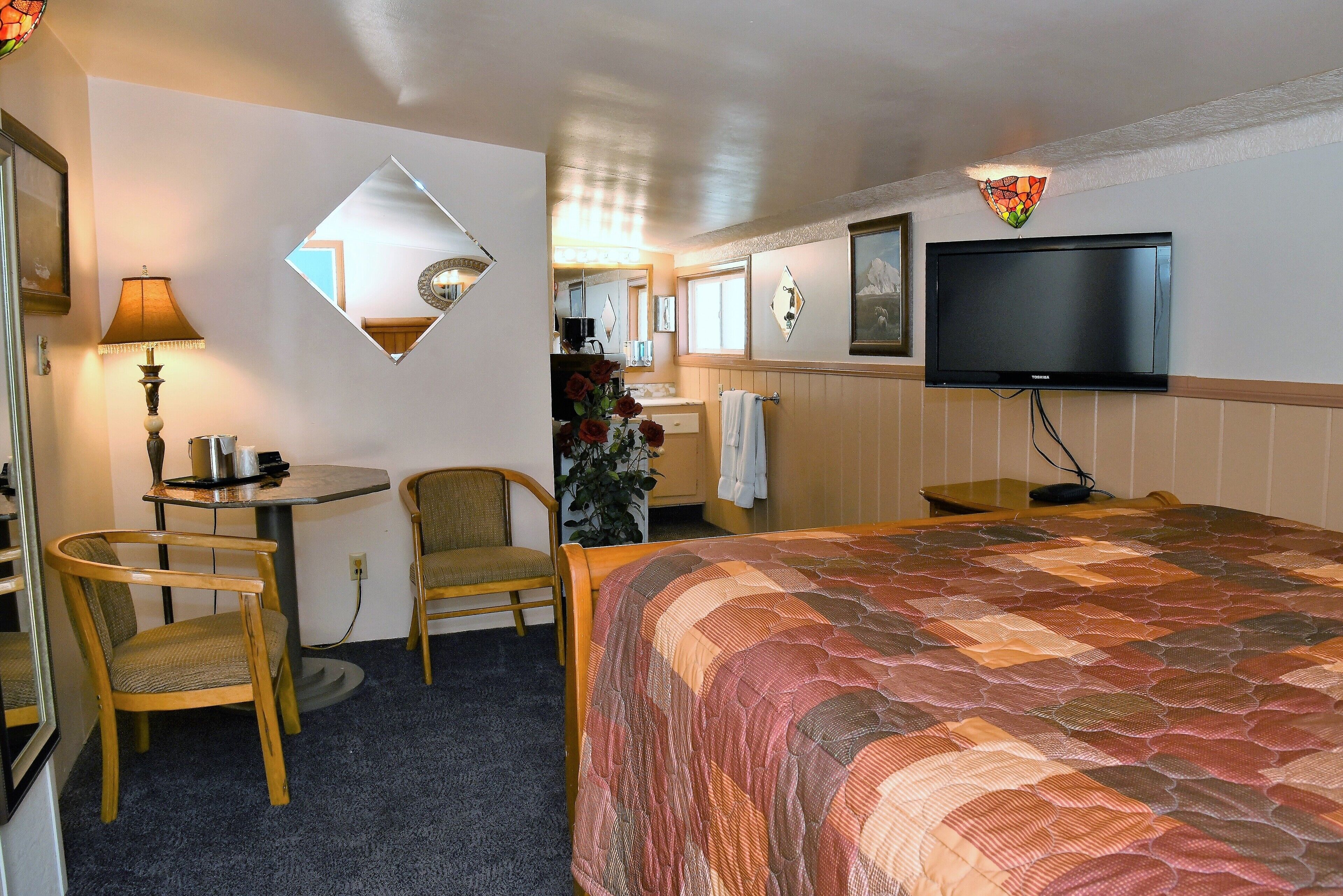 A hotel room with a bed, a desk with a lamp, two chairs, a television, and a mirror.