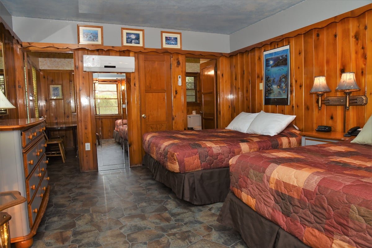 A room with two beds, a desk, and a dresser. There is a door leading to another room.