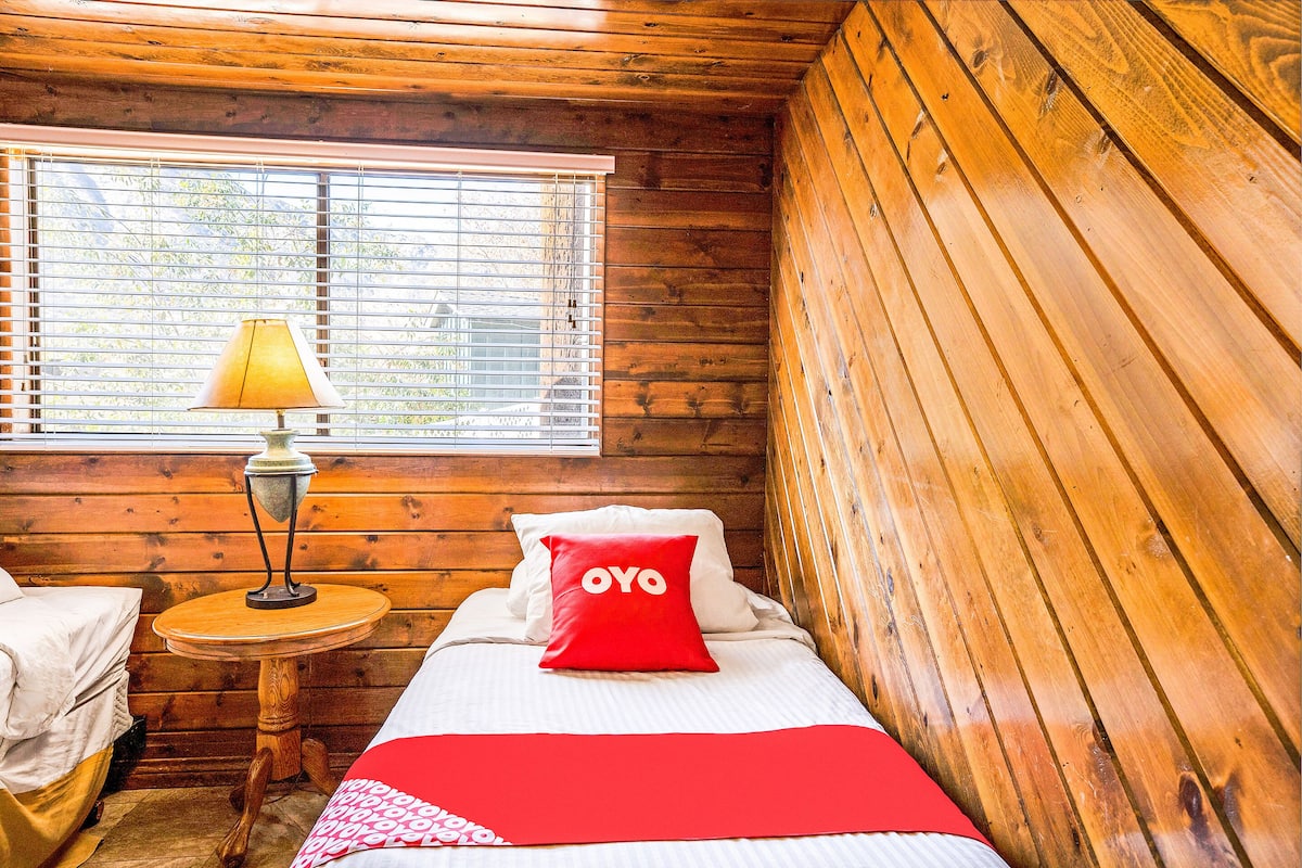 A wooden cabin room with a bed, a small table with a lamp, and a window with blinds.
