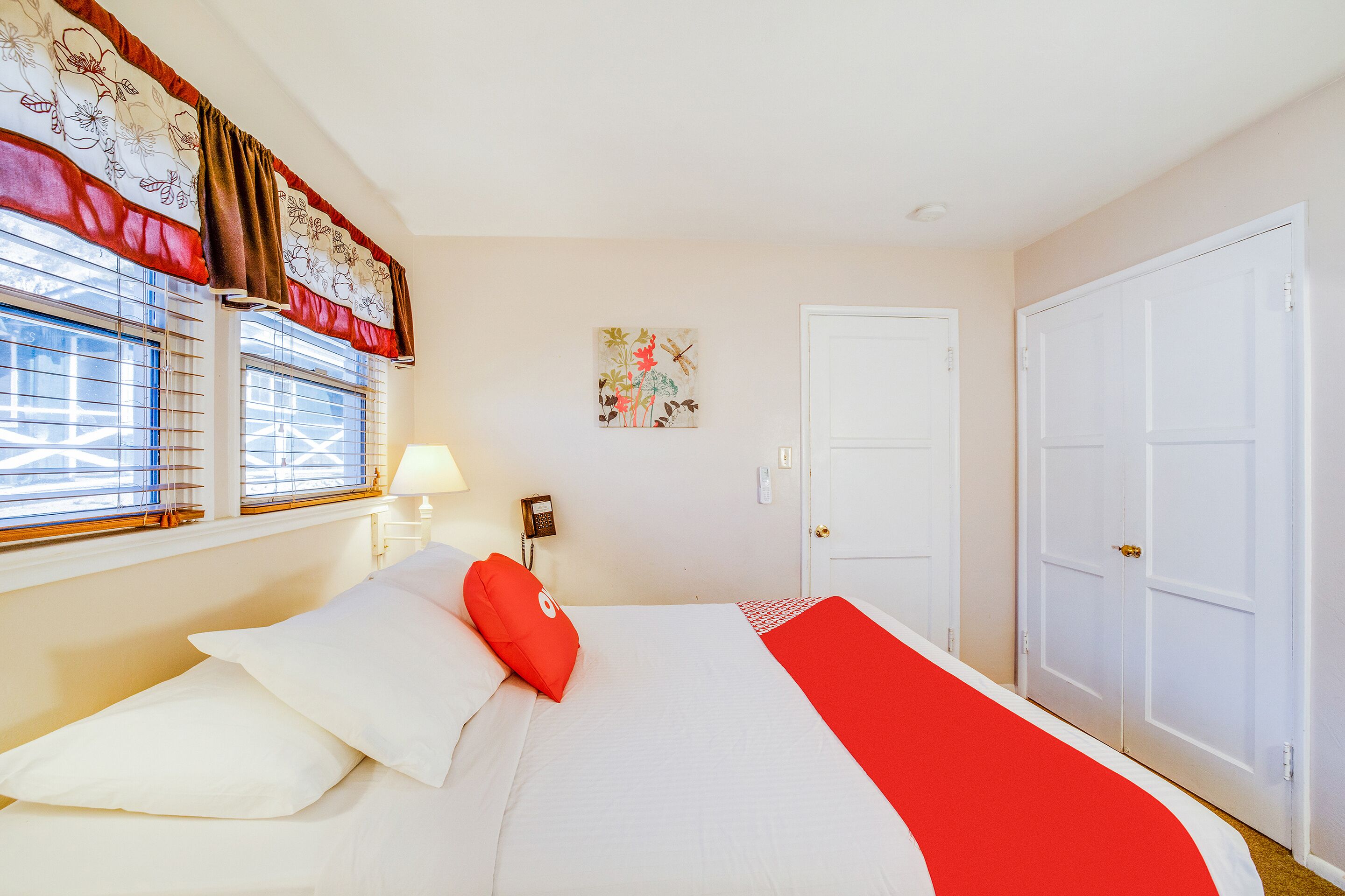 Un dormitorio con cama, paredes blancas, una ventana con cortinas, una puerta y una lámpara fijada a la pared.